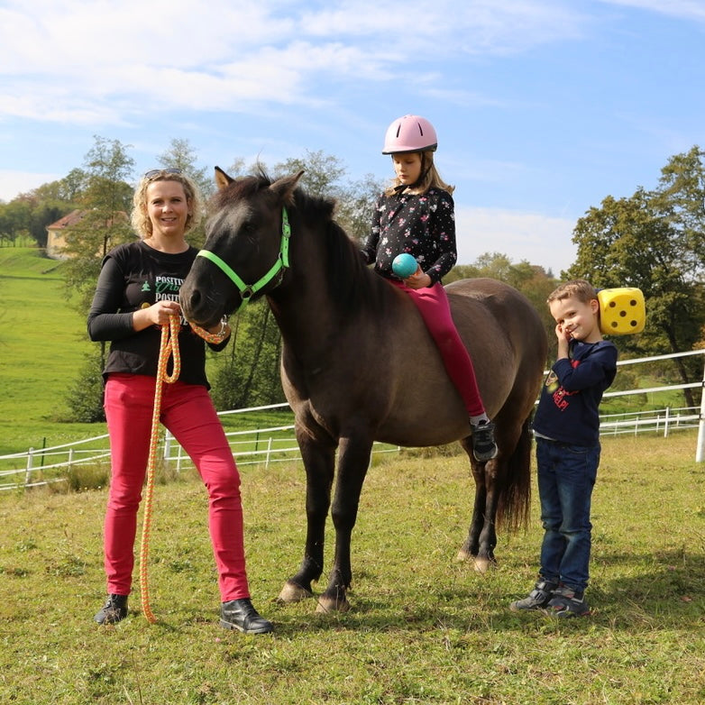 Frau hält ein braunes Pferd am Halfter, ein Mädchen sitzt auf dem Pferd und hält einen Ball in der Hand, ein Junge steht mit einem Schaumstoff Würfel neben dem Pferd
