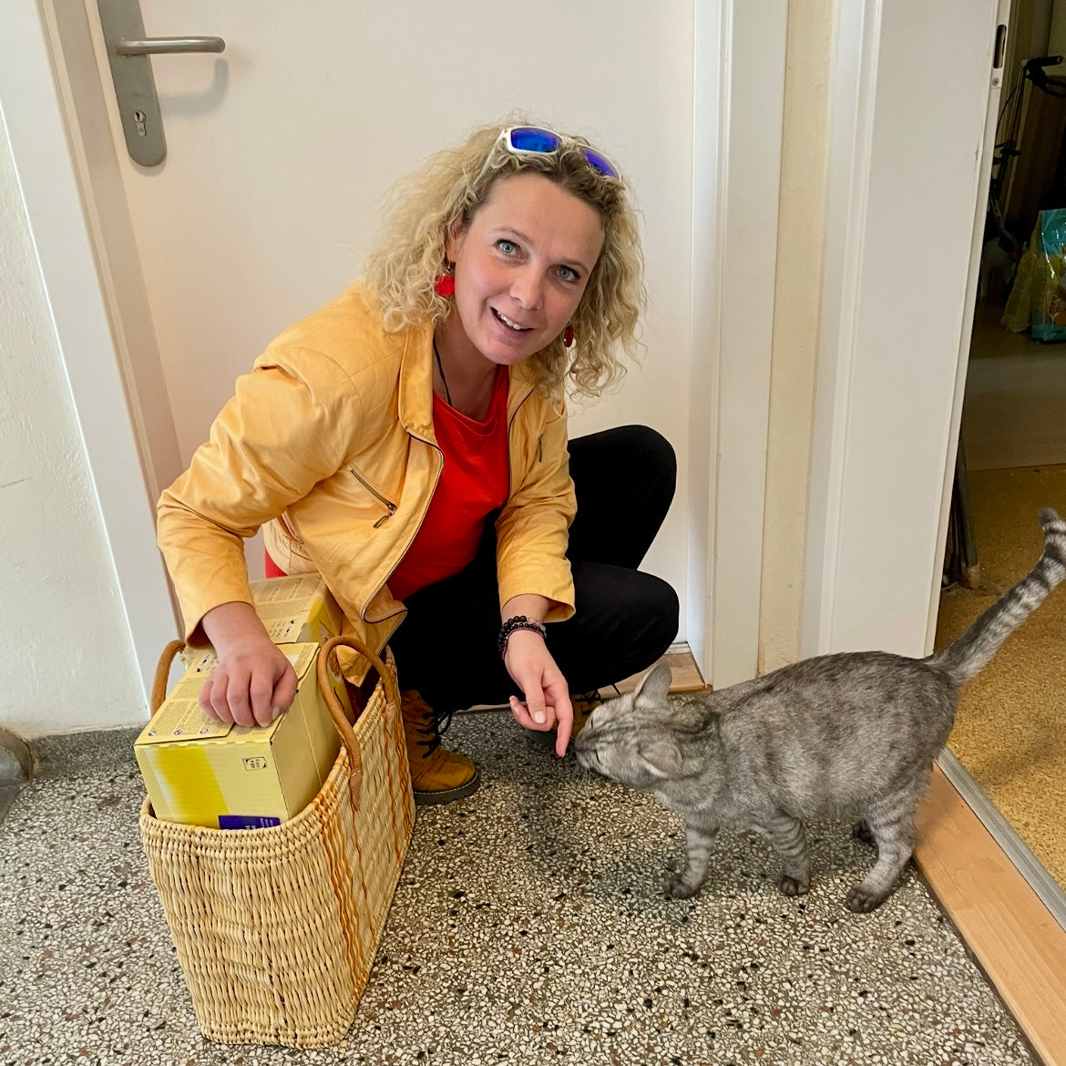 Frau hockt am Boden, Katze kommt zu ihr und riecht an ihrem Finger, daneben steht ein Korb mit Futter 